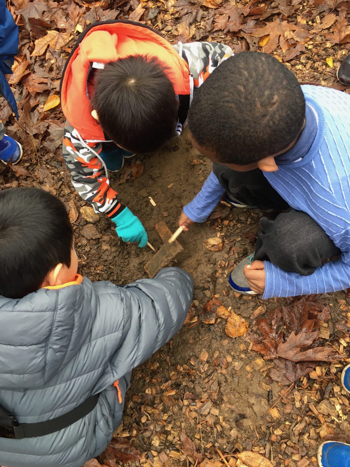 Three small children crouch to inspect an item in the muddy earth