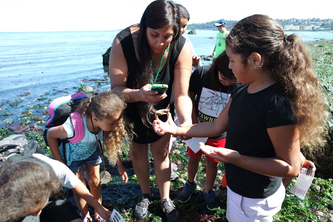 A Native woman takes a photo of a small crab held by Native child at the shore